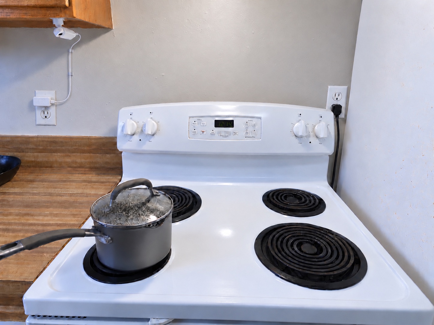 One StoveDetector unit mounted under the left cabinet beside an electric stove, with its power cord clipped down the wall to a nearby outlet on the left.